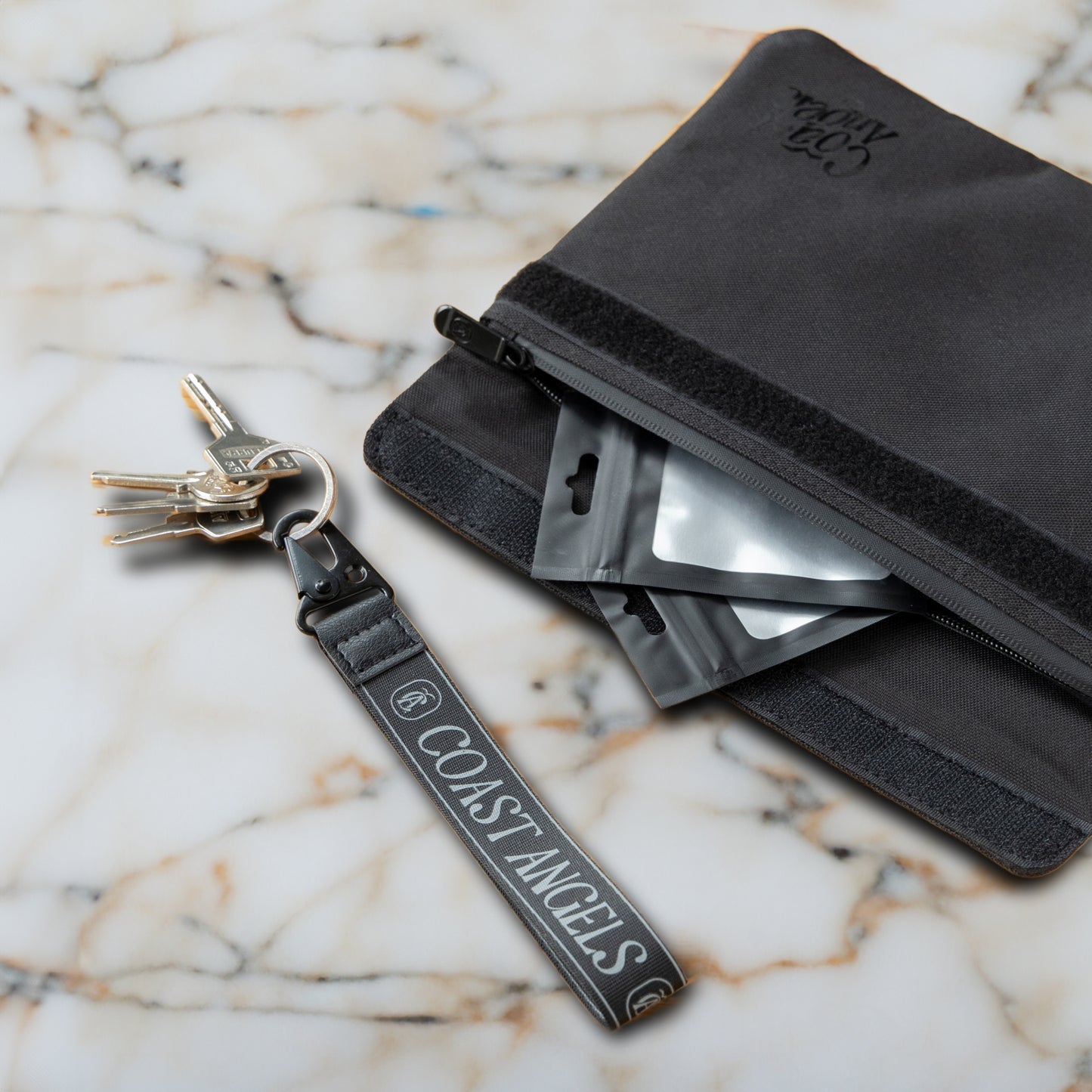 Black Coast Angels Soho smell-proof pouch shown partially unzipped on a marble surface, with resealable bags visible inside and a branded wrist strap holding a set of keys — showcasing portable, discreet functionality.