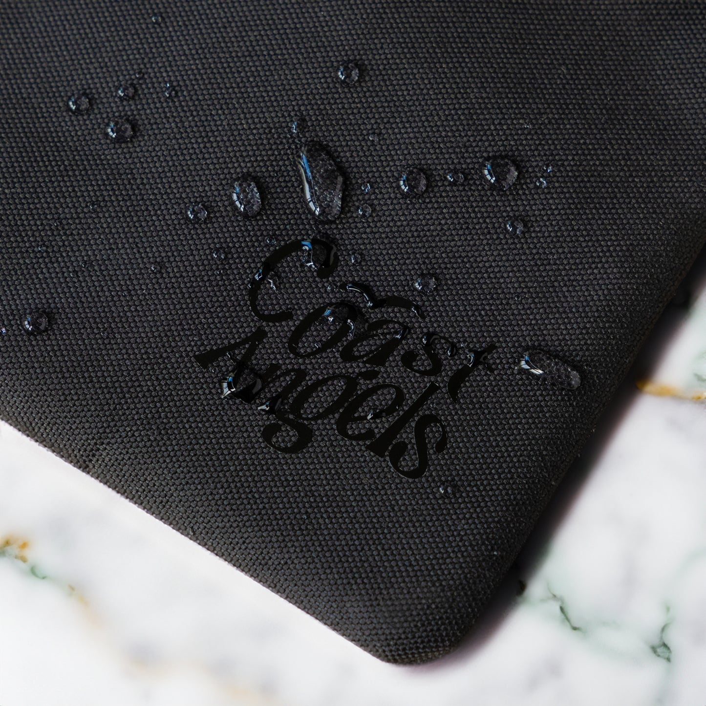 Close-up of the Coast Angels logo on a textured Soho black pouch, with water droplets beading on the surface — showcasing the bag’s water-resistant material and durable finish.