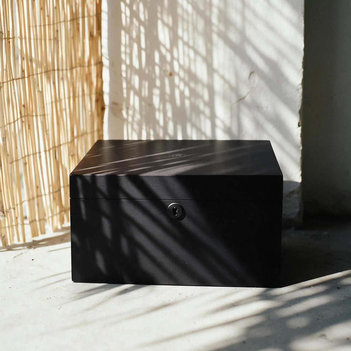 A matte black wooden storage box with a small round lock, placed on a concrete floor in natural light with bamboo and window shadows cast across it.