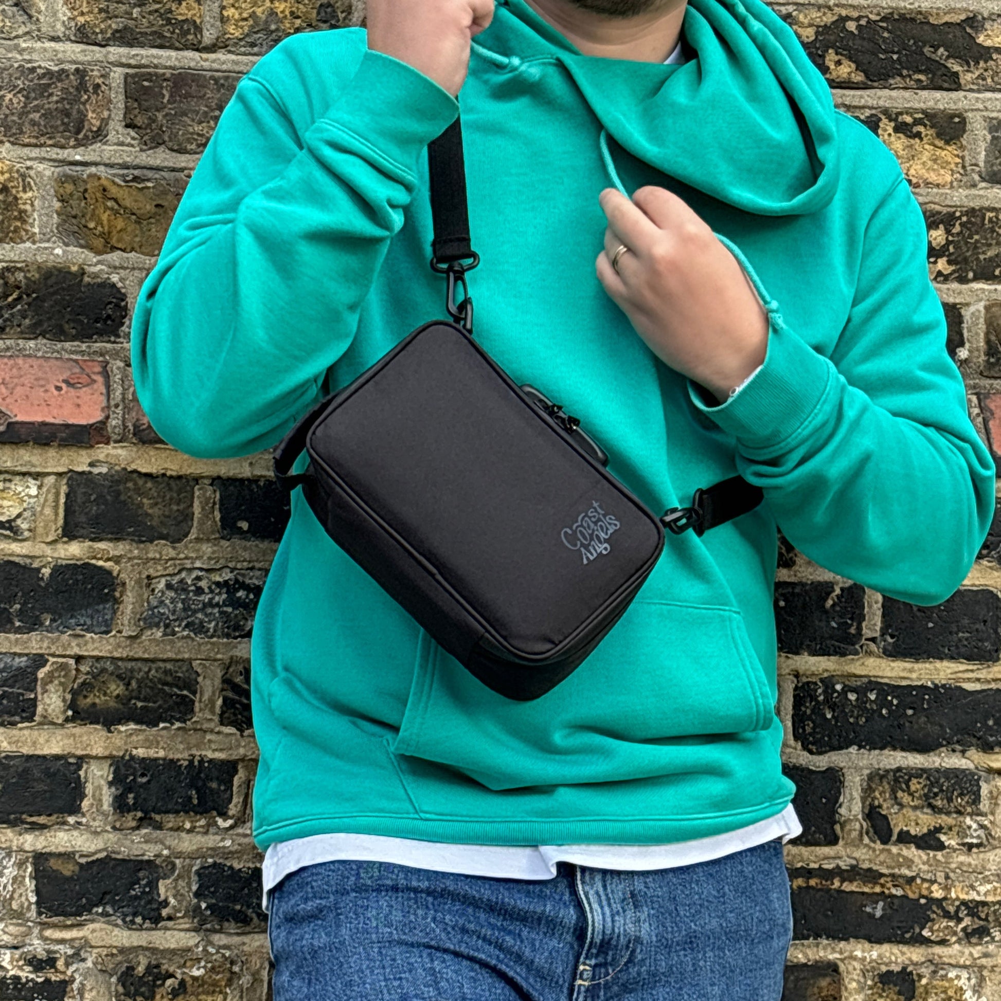 Man wearing a turquoise hoodie with the Coast Angels Dalston Bag against a brick wall, highlighting the bag's sleek and versatile style.