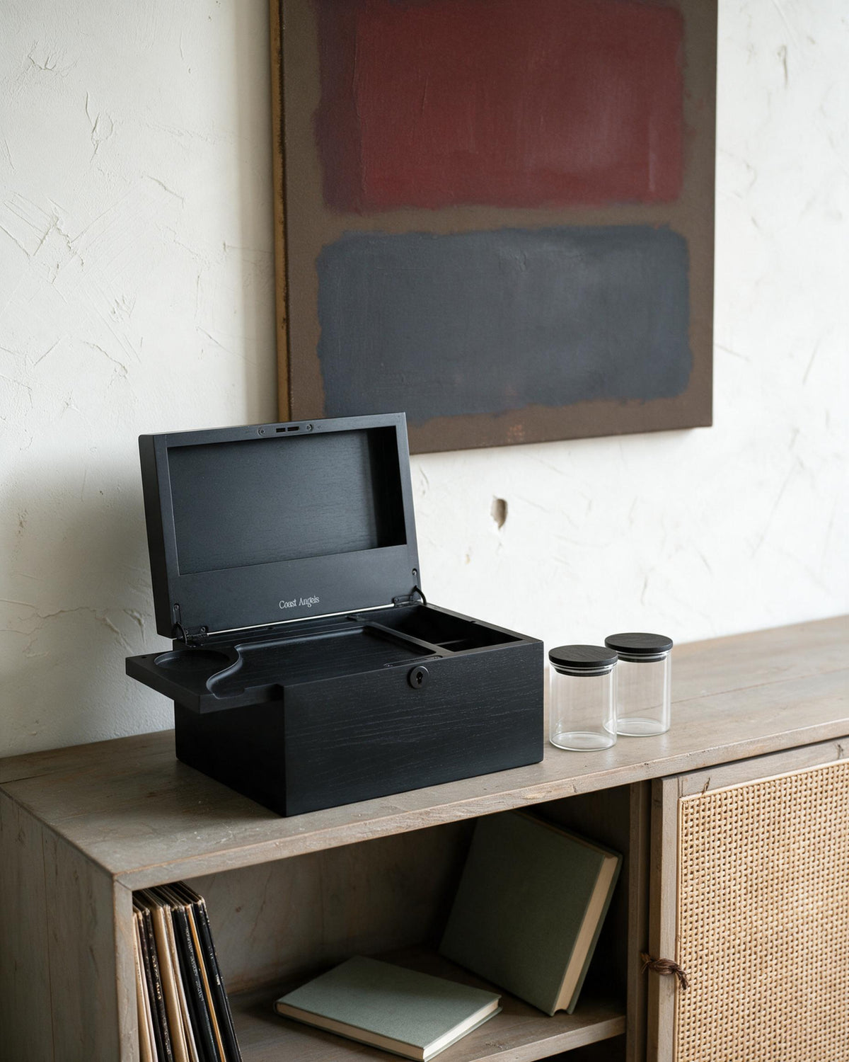 A Coast Angels black wooden storage box with its lid open, placed on a light wood cabinet in a styled interior setting with two clear glass jars nearby.