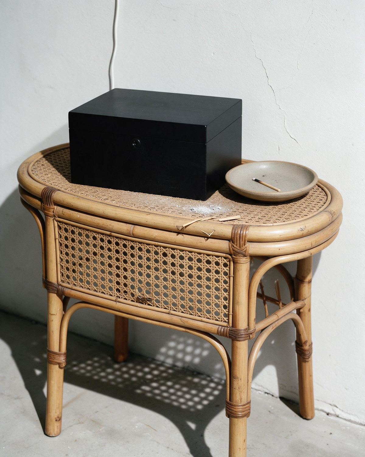 A natural rattan side table with a woven cane front and bamboo legs, styled with a black wooden box and a small ceramic dish against a white wall.