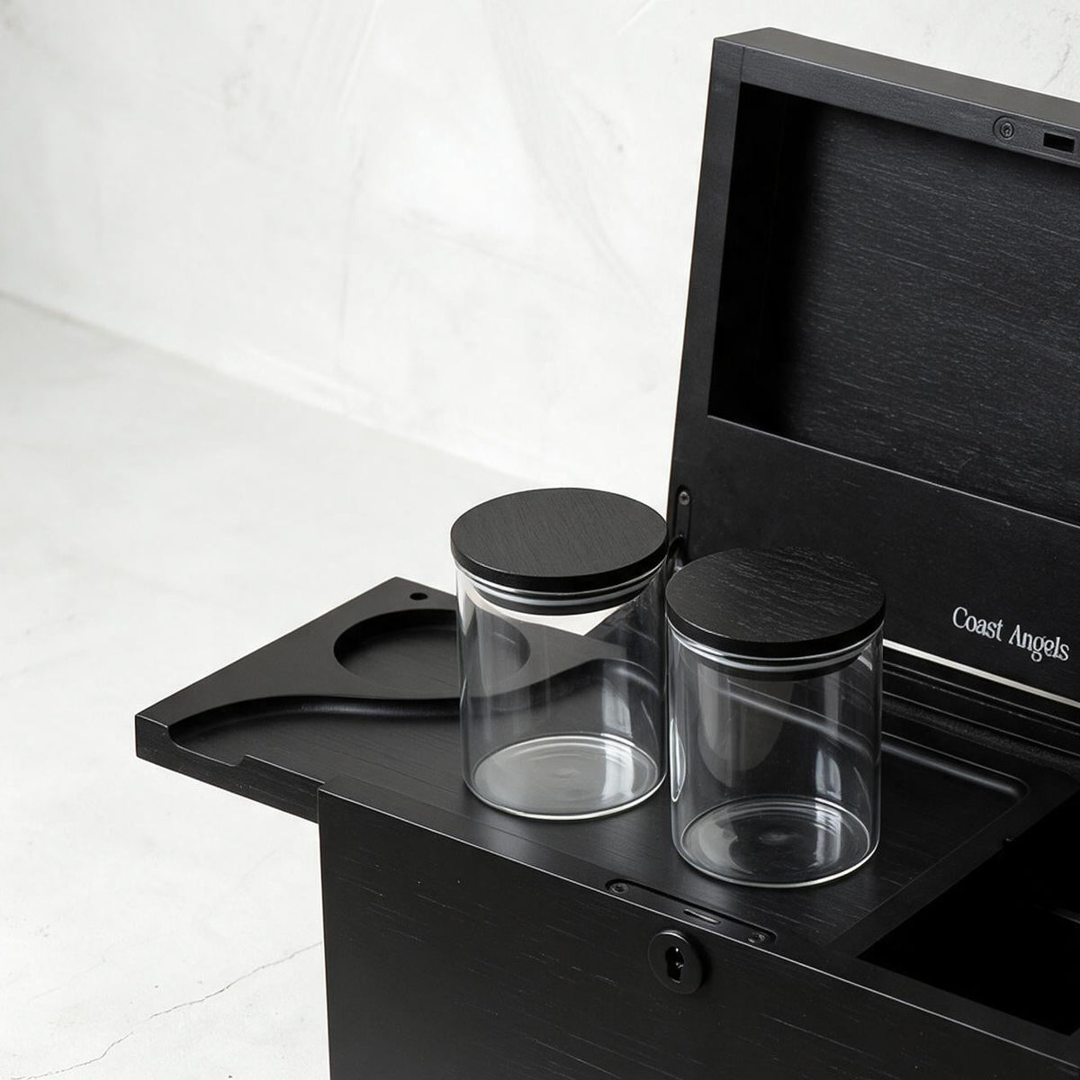 A Coast Angels black wooden storage box with its lid open, holding two clear glass jars with black lids and a black utensil, photographed on a light stone surface against a textured white wall.