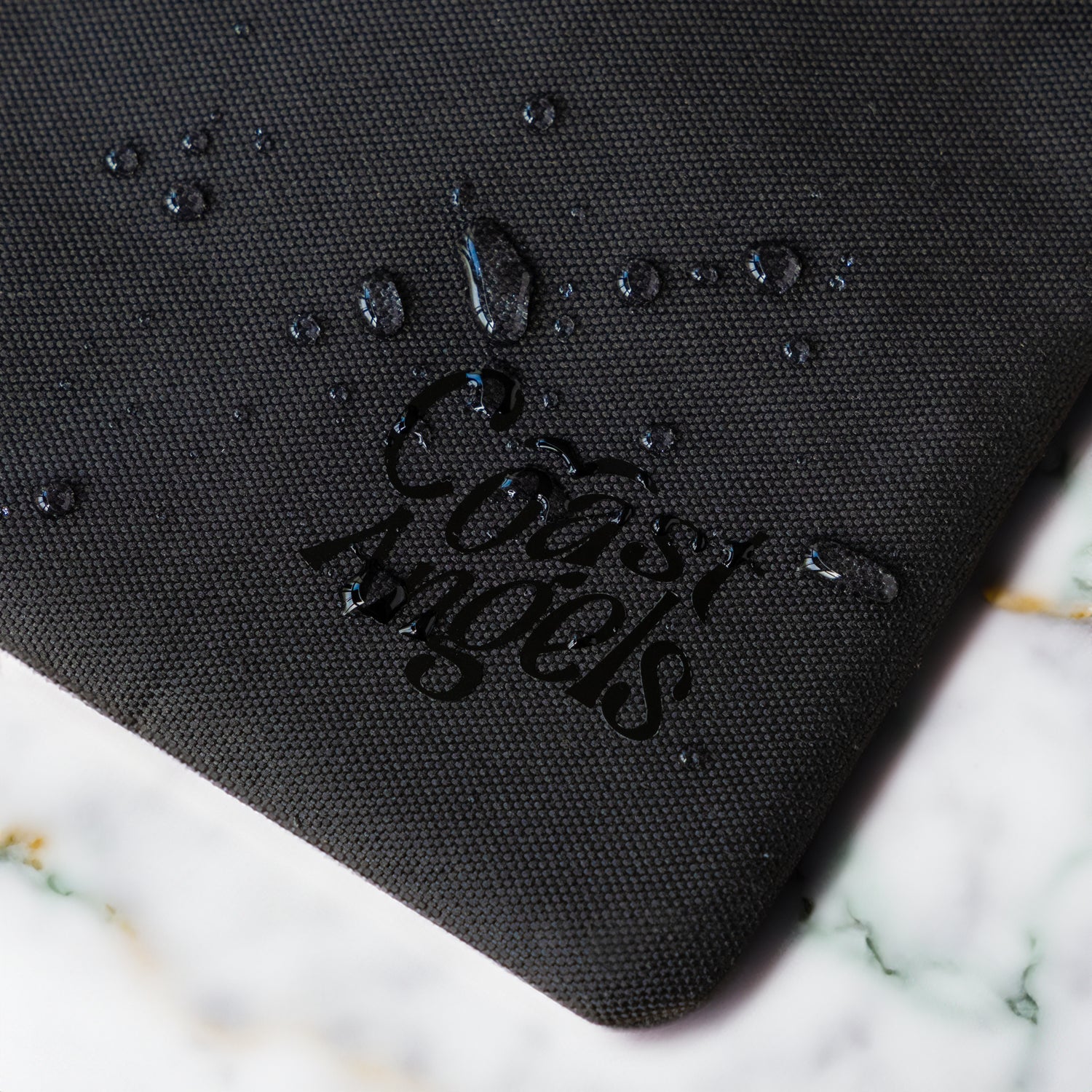 Close-up of the Coast Angels logo on a textured Soho black pouch, with water droplets beading on the surface — showcasing the bag’s water-resistant material and durable finish.