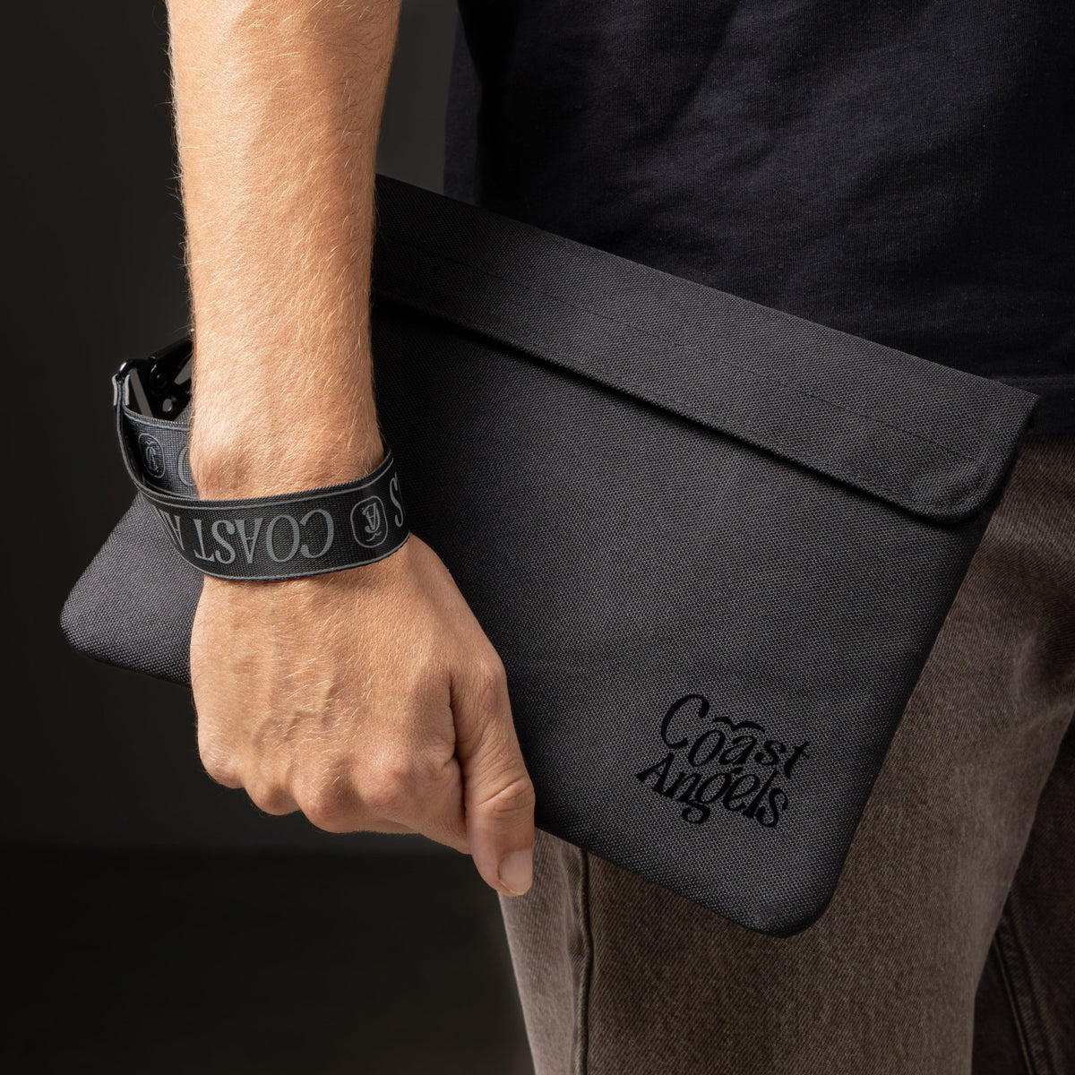 Person holding the black Coast Angels Soho pouch by their side, with the branded wrist strap wrapped around their wrist — showcasing the pouch’s portability, sleek design, and everyday wearability.
